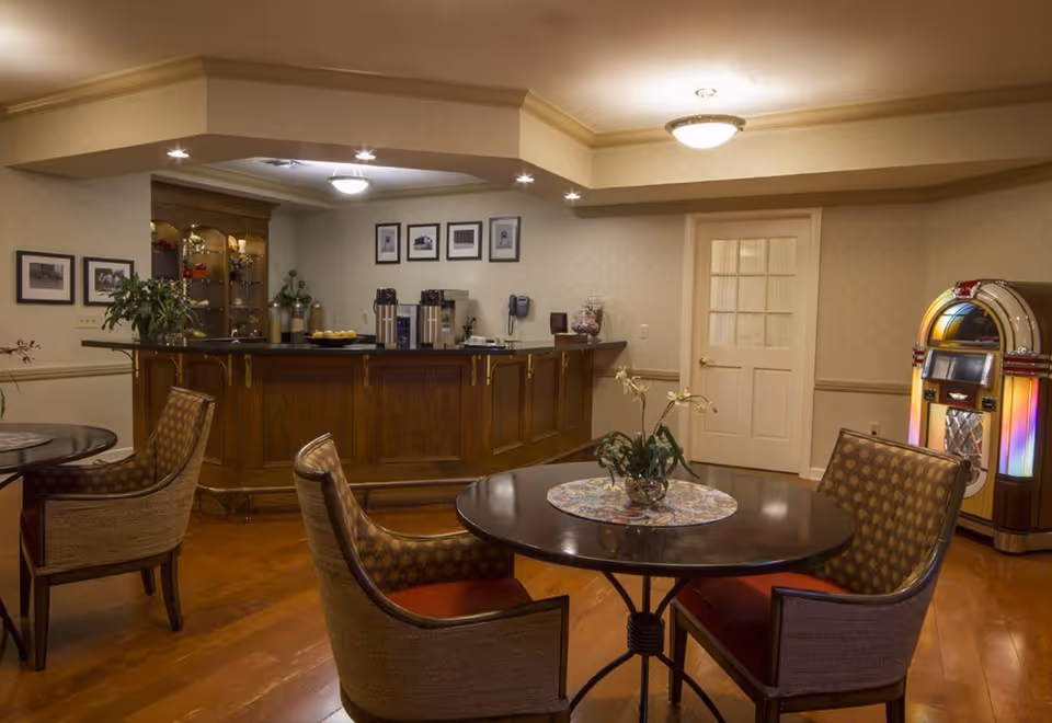 A cozy interior room with wooden flooring featuring a round table with four cushioned chairs around it. A small plant centerpiece is on the table. In the background, there is a wooden counter with coffee machines and framed pictures on the wall above it. To the right, there is a vintage jukebox with colorful lights. The room has warm lighting and a door with glass panels.