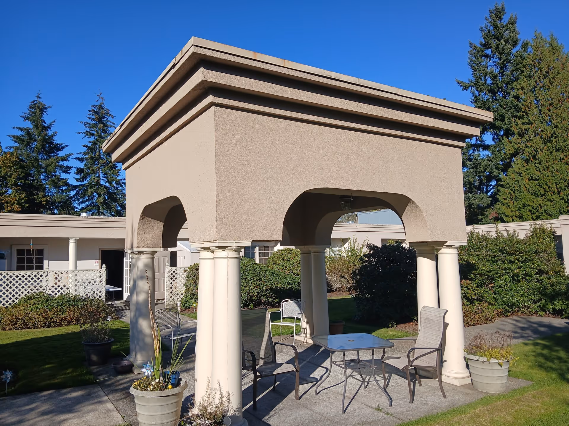 Outdoor seating area with a beige gazebo supported by four white columns, surrounded by greenery and shrubs, with a clear blue sky overhead. There are four metal chairs and a glass-top table under the gazebo, and potted plants around the structure.