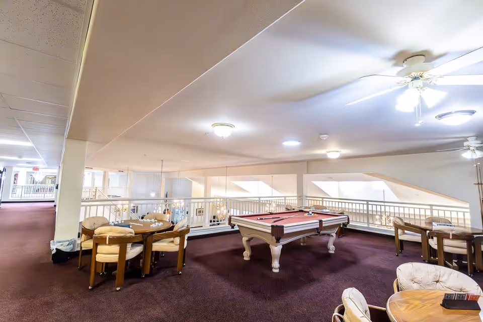 Interior view of a senior living facility's recreational area featuring a pool table with a red felt top, surrounded by several round tables with cushioned chairs. The space has a carpeted floor, ceiling fans with lights, and a railing overlooking a lower level.
