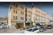 Four-story beige brick nursing facility with green entry awnings, parked cars along the street, and overhead utility wires.