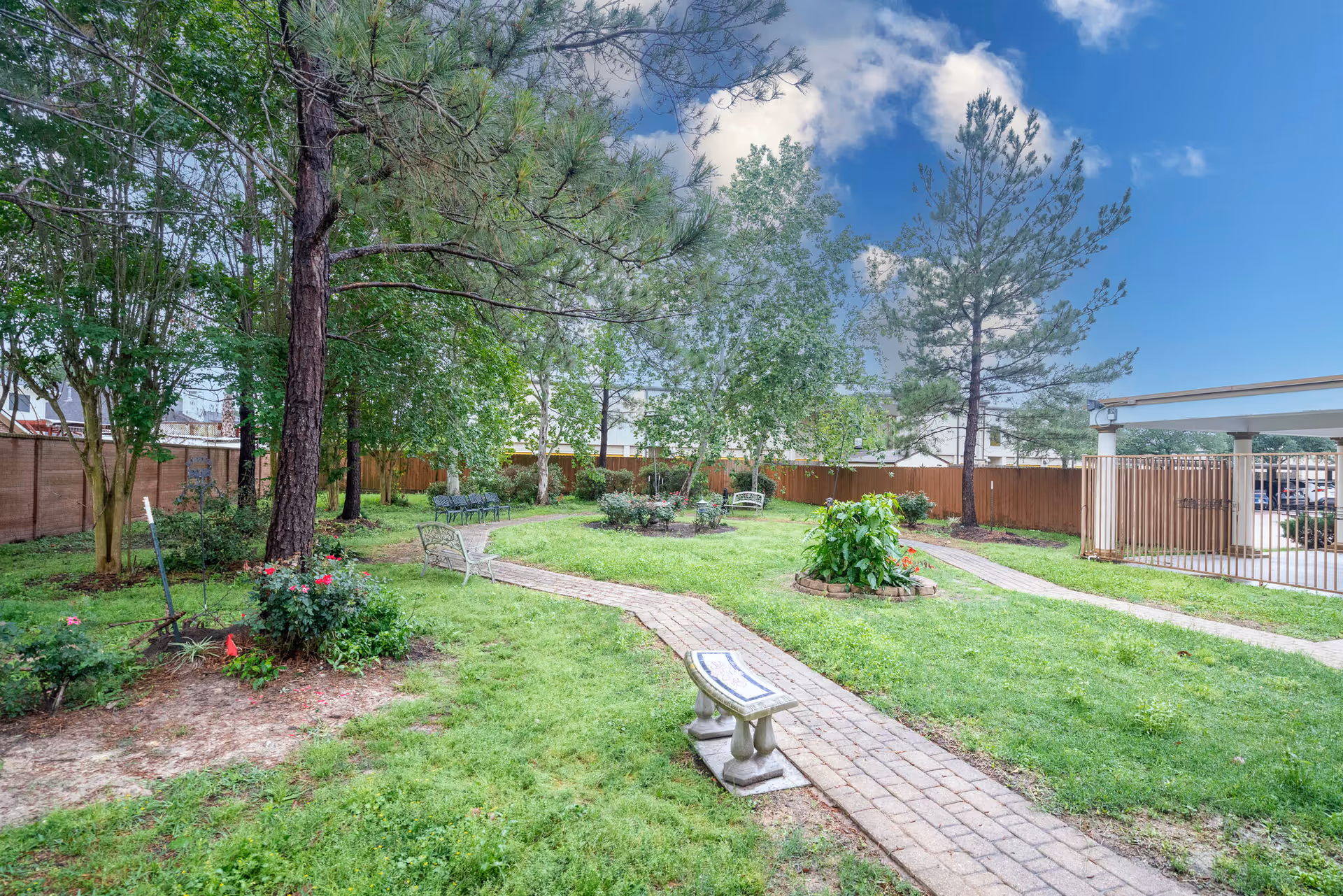 A peaceful outdoor garden area with green grass, trees, flower beds, and several benches along a winding brick pathway. A wooden fence encloses the garden, and a covered parking area is visible in the background under a partly cloudy blue sky.