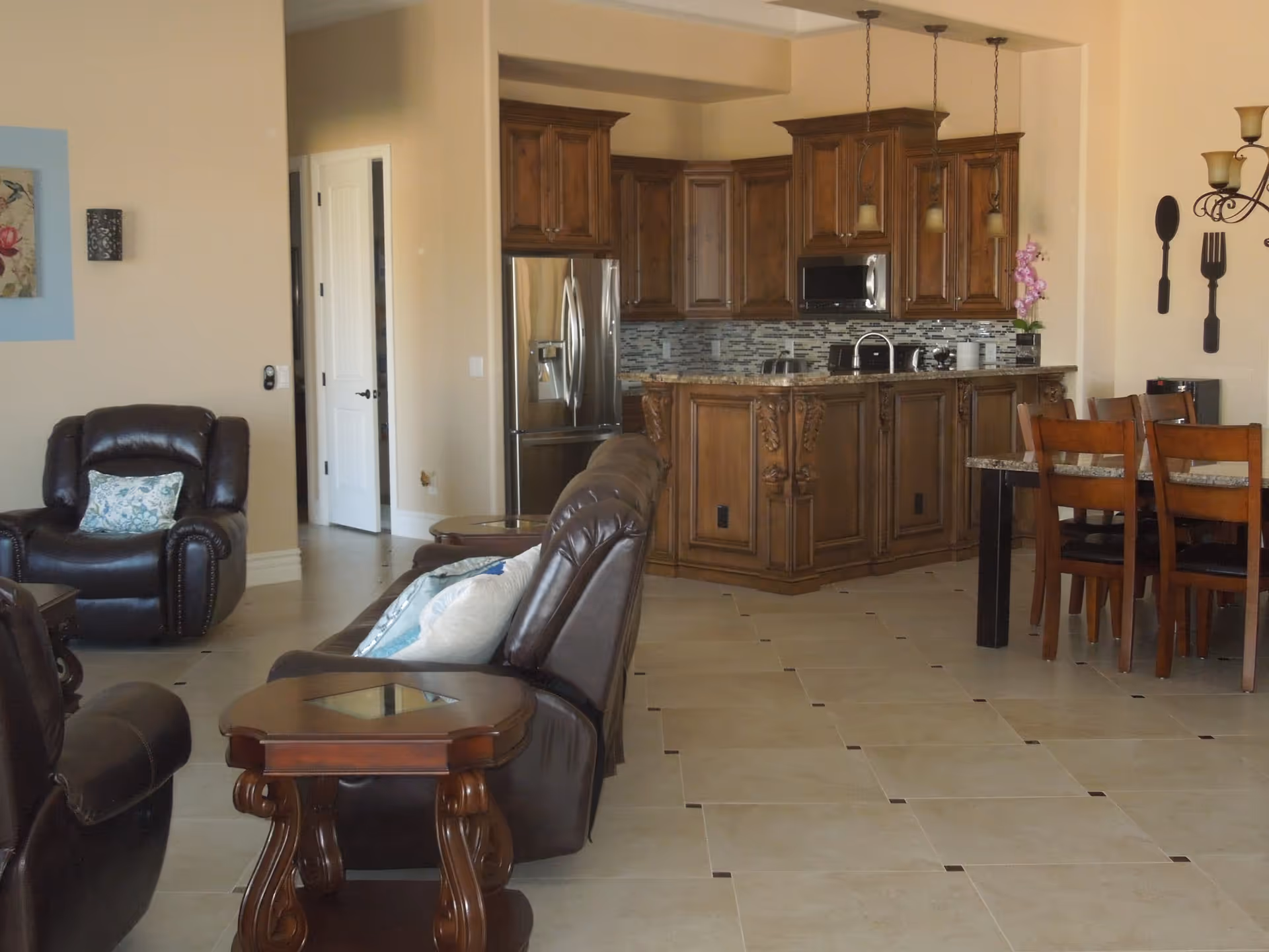 Open interior space featuring a living room with dark leather armchairs and a wooden side table, adjacent to a kitchen with wooden cabinetry, stainless steel refrigerator, and a kitchen island with granite countertop. A dining area with wooden chairs and a table is visible on the right side, with wall decorations including large spoon and fork silhouettes.