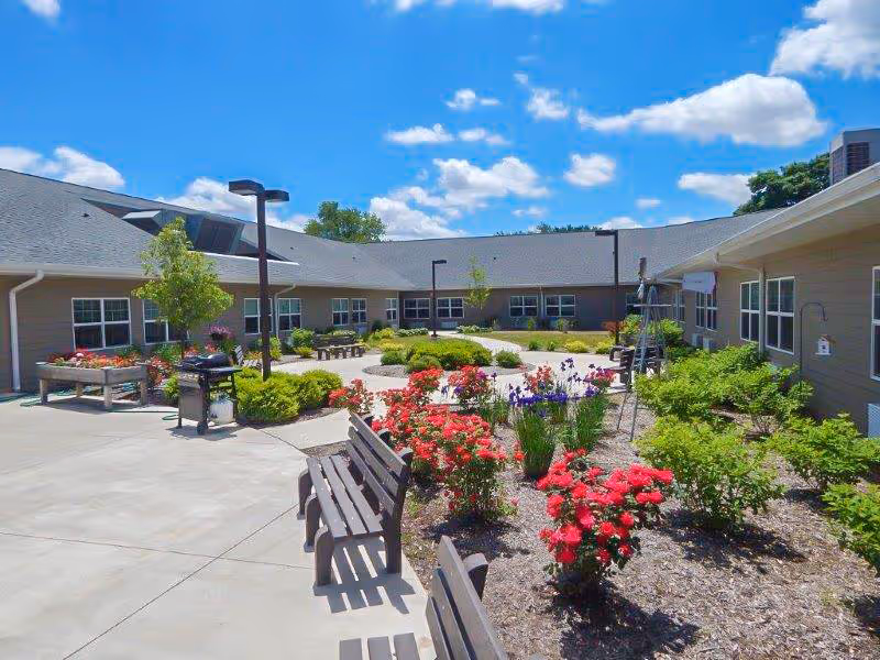 Sunlit courtyard with benches, flower beds, and walkways surrounded by single-story care facility buildings under a blue sky.