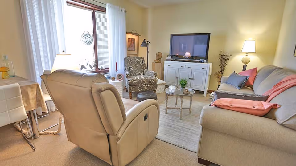 A cozy living room with a beige recliner chair, a patterned armchair, a beige sofa with colorful pillows, a small round coffee table, a white TV stand with a flat-screen TV, and soft lighting from lamps. There is a window with white curtains and a small dining table with chairs in the corner.