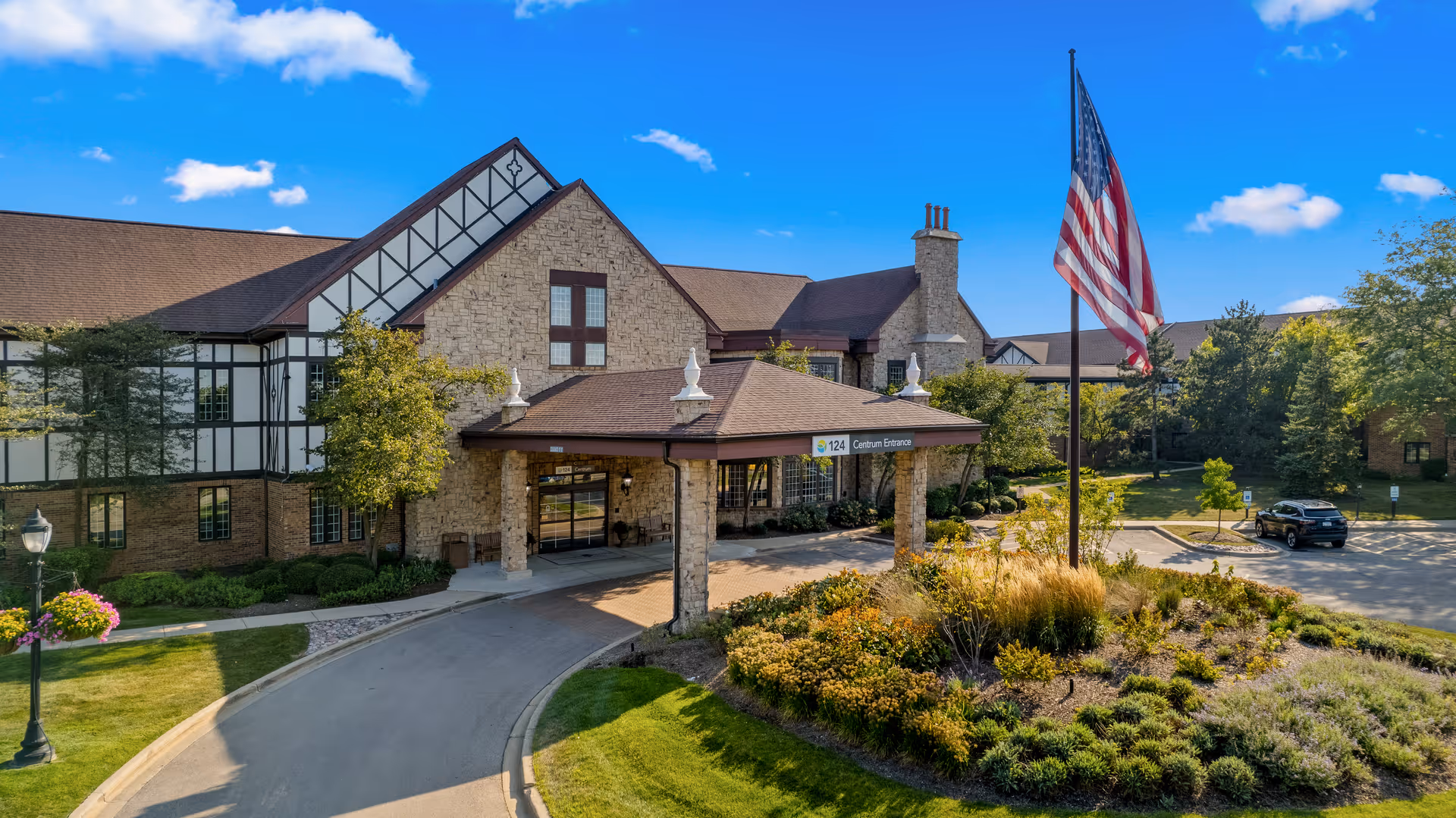 Exterior view of a senior living facility building with stone and timber facade, a covered entrance with a sign reading '124 Centrum Entrance,' a landscaped roundabout with plants and an American flag on a flagpole, and a clear blue sky with some clouds.