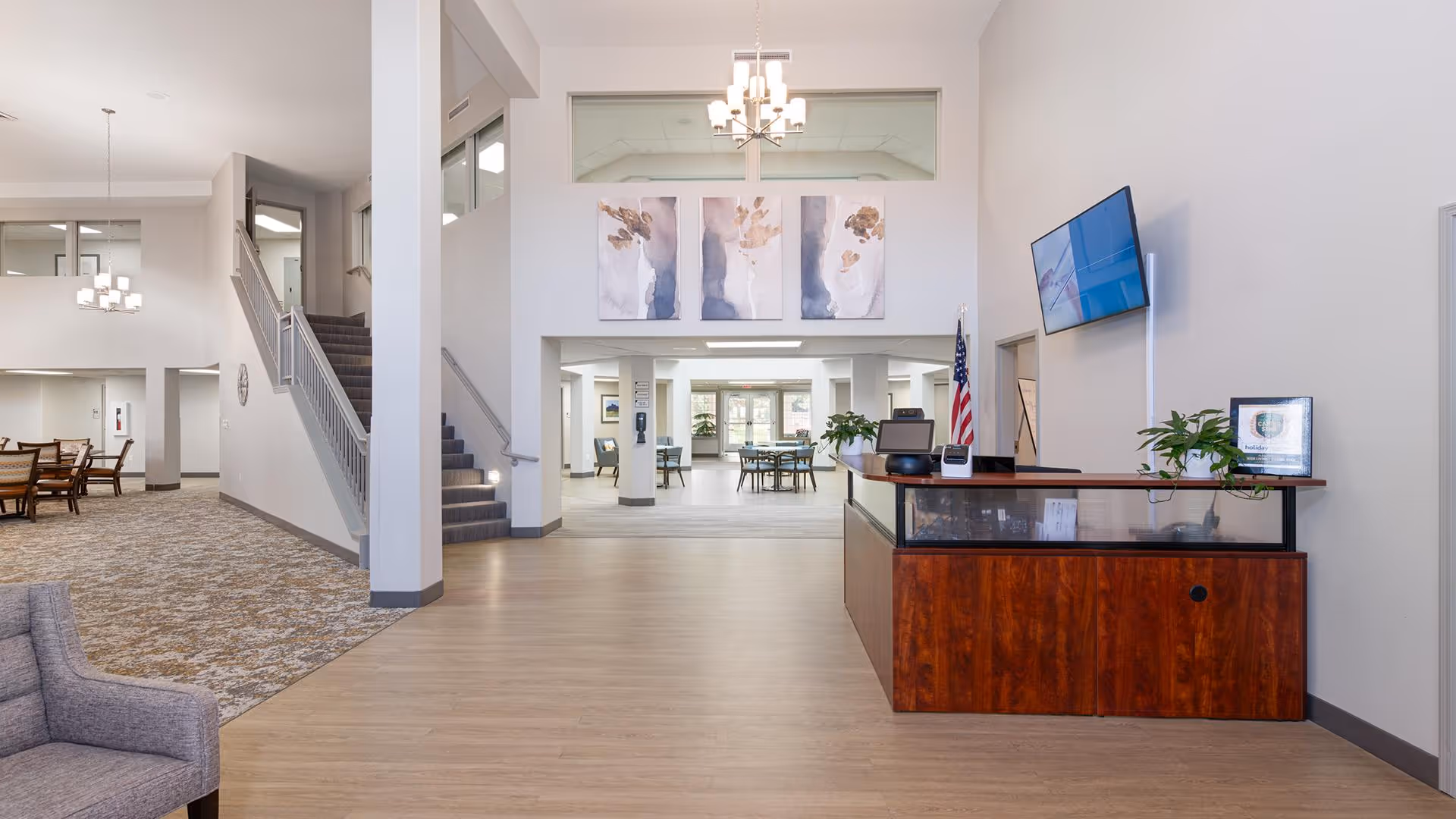 Interior view of a senior living facility lobby area with a wooden reception desk on the right, a mounted TV above it, and an American flag behind. There is a staircase leading upstairs on the left, a dining area with tables and chairs in the background, and modern light fixtures hanging from the ceiling. The space is bright with neutral colors and decorative artwork on the wall above the entrance to another room.