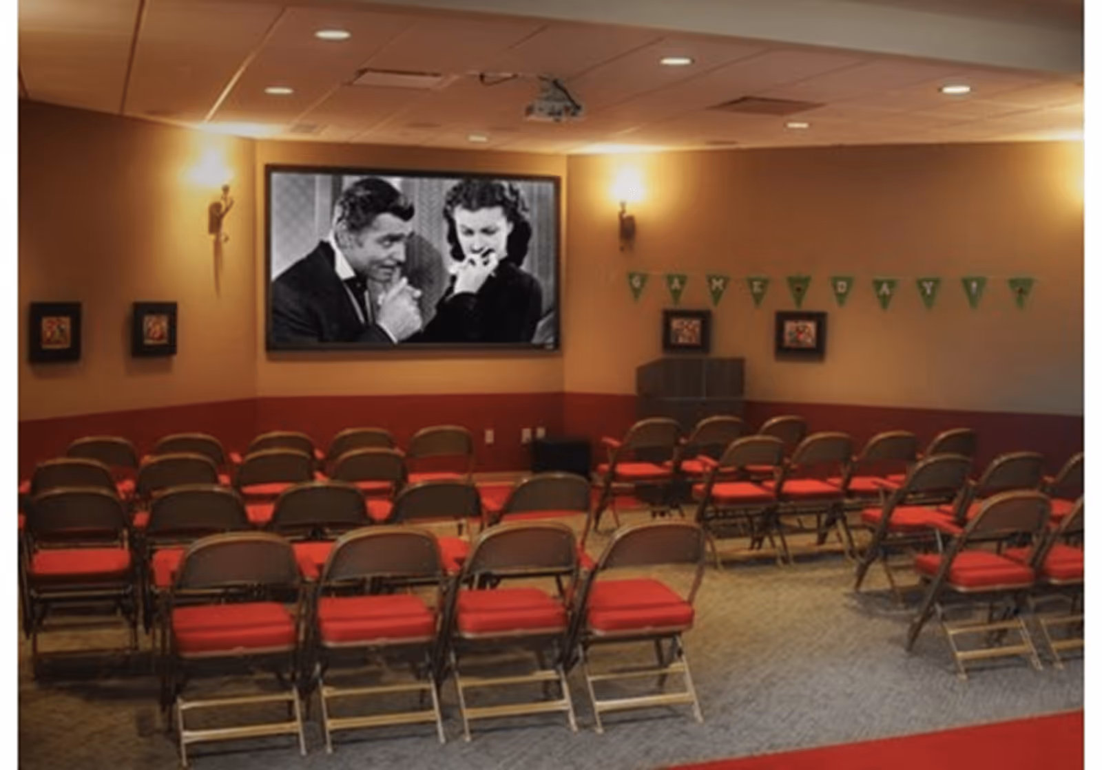 A small theater room with rows of folding chairs with red cushions facing a large screen showing a black and white movie. The room has warm lighting with wall sconces and a banner on the wall spelling 'CINEMATOPIA'.