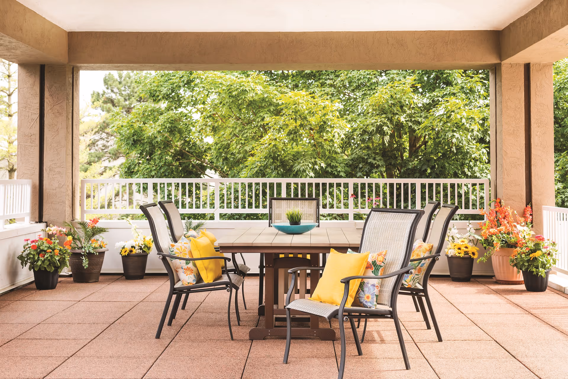 Outdoor covered patio area with a rectangular wooden table surrounded by six metal chairs with cushions. The patio is decorated with multiple potted plants and flowers, and green trees are visible beyond the white railing.