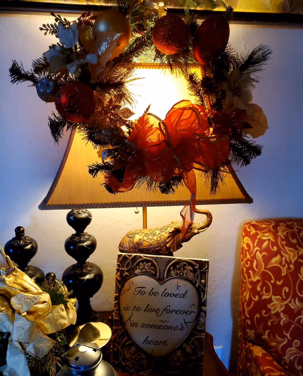 Table lamp decorated with a festive wreath and ribbon, sitting on a side table with ornaments, a heart-shaped plaque and a patterned armchair beside it.