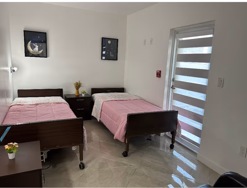 Two single beds with pink covers in a bright, minimally decorated bedroom with a nightstand and a door with striped blinds.