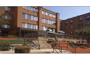 Exterior view of a multi-story brick building with large windows, ramps, and stairs leading to the entrance. The area around the building includes landscaped bushes and red mulch beds under a clear blue sky.