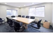 Small conference room with a rectangular table surrounded by black chairs and two windows letting in daylight.