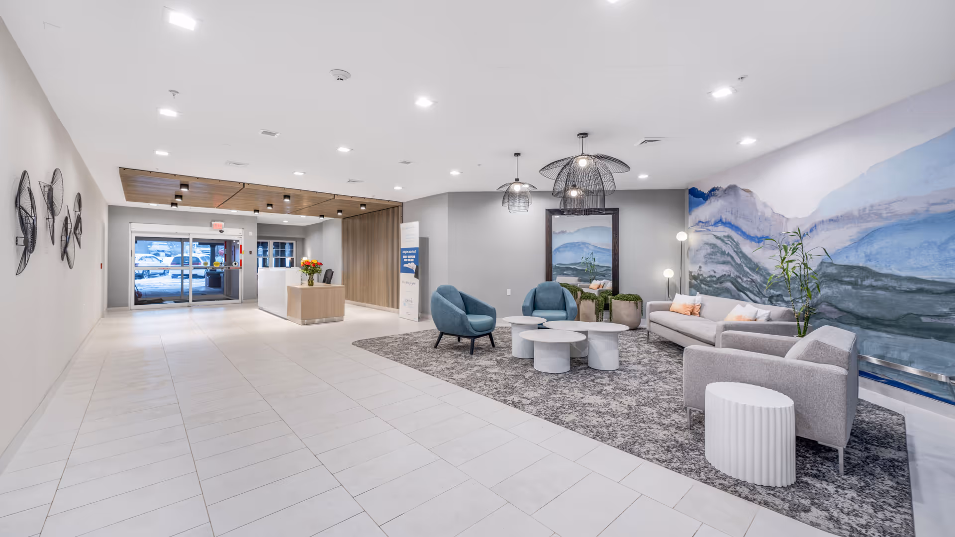 A spacious and modern reception area with a seating lounge. The lounge features a gray carpet, two gray sofas, two blue armchairs, three round white coffee tables, and a tall plant. The wall behind the seating area has a large mural with blue and gray abstract mountain designs. Pendant lights hang from the ceiling above the seating area. The reception desk is visible in the background near the entrance with glass doors, and there are decorative wall art pieces on the left wall.