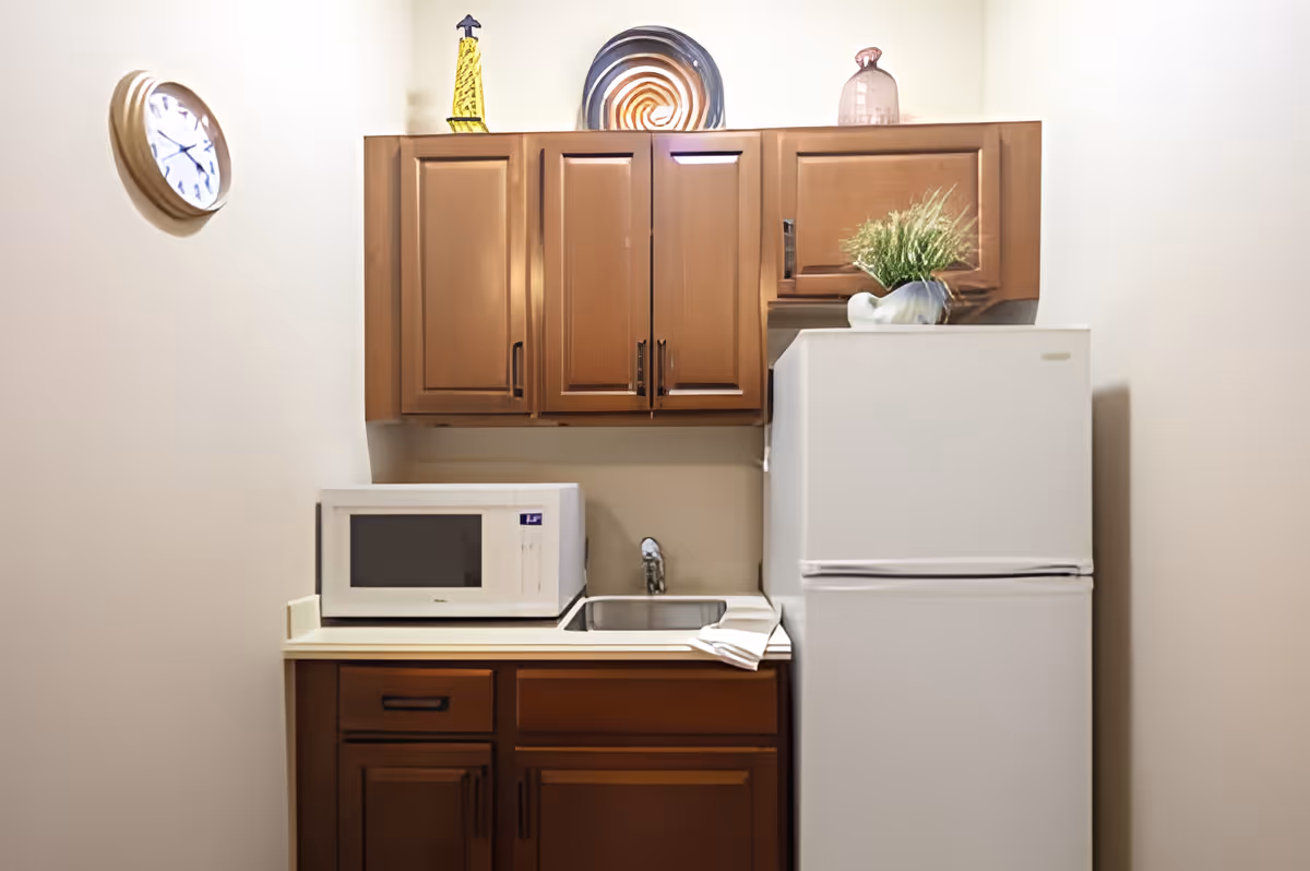 Small kitchen area with wooden cabinets, a white microwave on the countertop, a stainless steel sink with a faucet, and a white refrigerator. Decorative items including a yellow lighthouse figurine, a spiral plate, a pink glass bottle, and a small plant in a white vase are placed on top of the cabinets and refrigerator. A wall clock is mounted on the left wall.