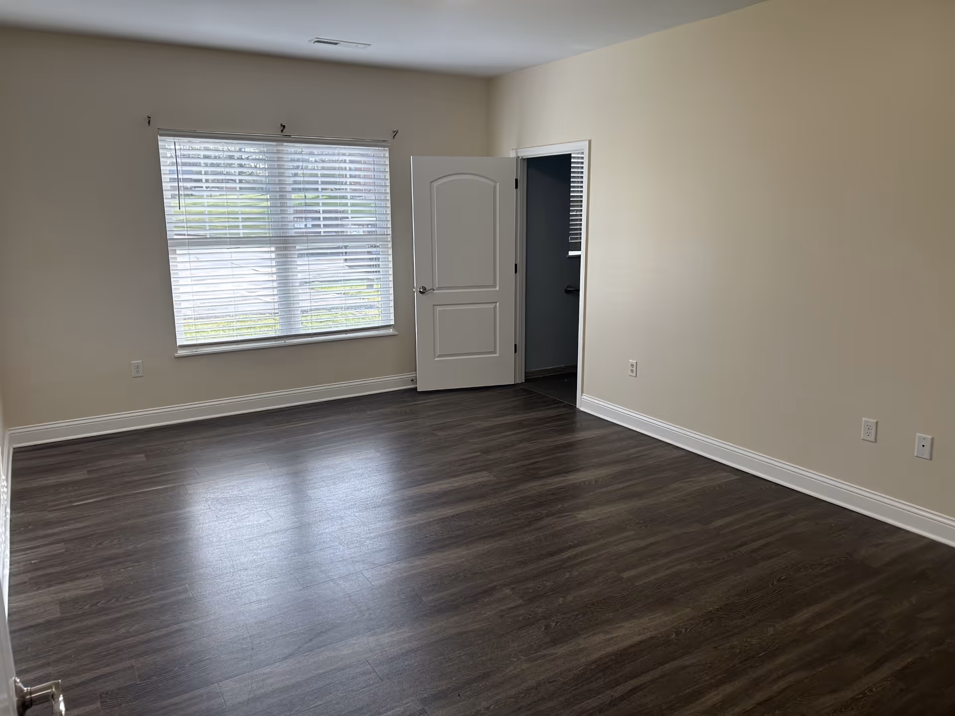 Empty room with dark wood flooring, beige walls, a large window with white blinds, and a white door slightly open leading to another room.