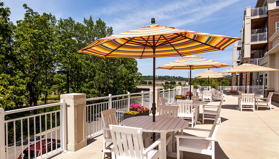 Outdoor patio with striped umbrellas, round tables and white chairs along a railing overlooking trees and a building.