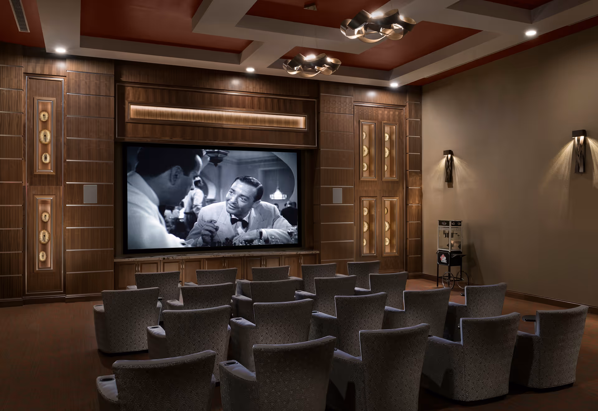 A cozy movie theater room with multiple rows of cushioned armchairs facing a large screen showing a black-and-white film. The walls are decorated with wood paneling and soft lighting fixtures, and there is a popcorn machine in the corner.