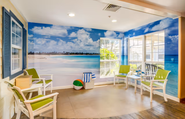 Common seating area with white chairs with green cushions, small tables and a large tropical beach mural on the walls.