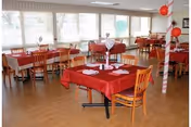 A dining room with several wooden tables and chairs arranged neatly. The tables are covered with red tablecloths and decorated with folded napkins and centerpieces. Large windows along one wall allow natural light to fill the room.