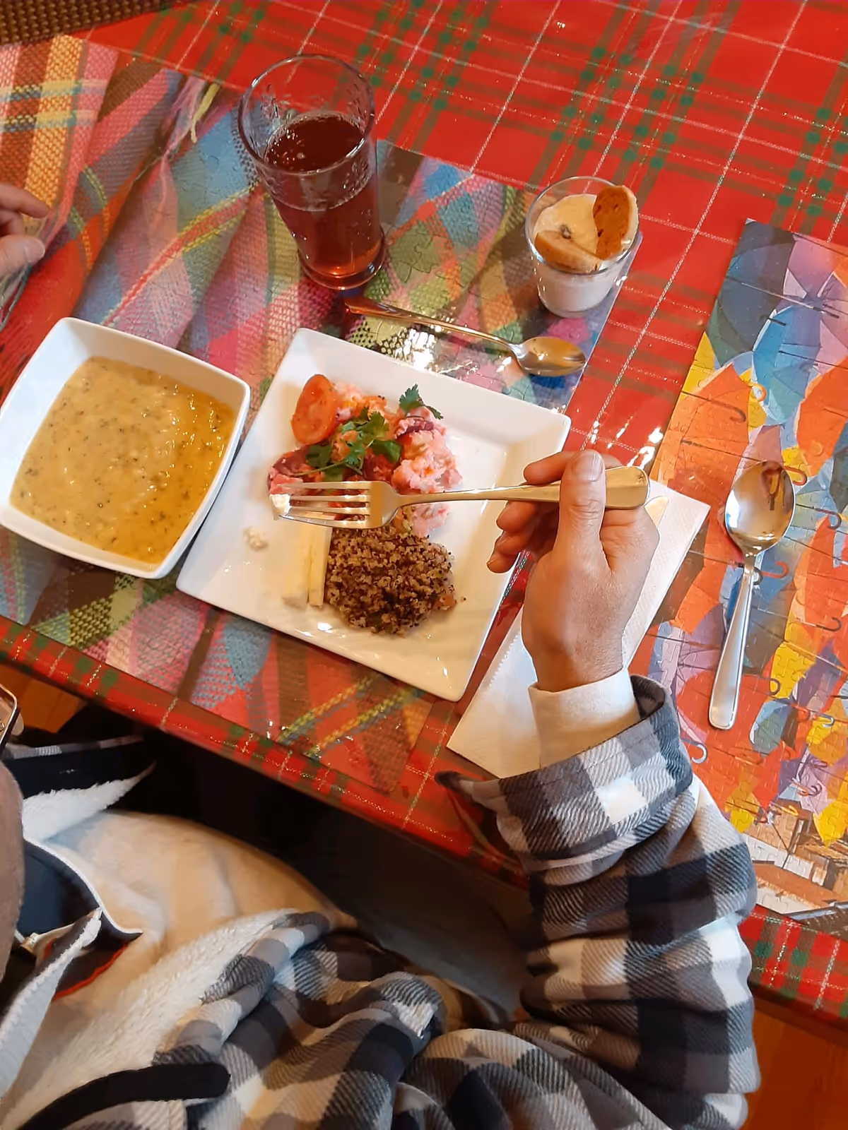 A person in a plaid jacket holding a fork over a plate of food with a bowl of soup, a glass of drink and a small dessert on a patterned tablecloth.
