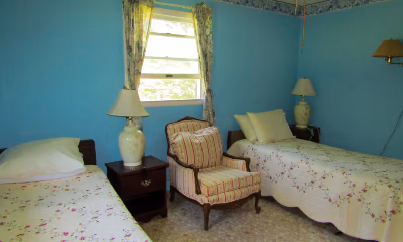 A small bedroom with two single beds covered in floral bedspreads, a striped upholstered armchair between them, two bedside tables each with a lamp, and a window with floral curtains letting in natural light. The walls are painted blue.