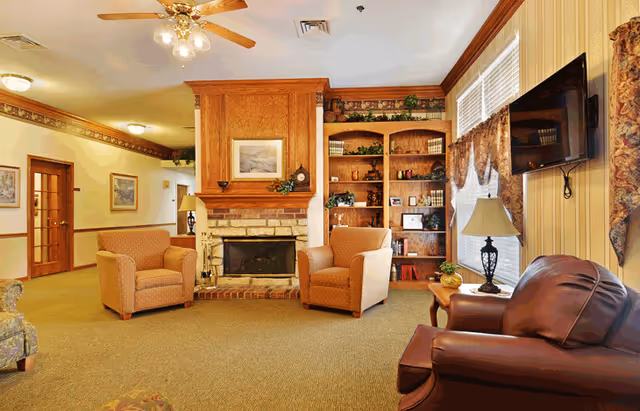 Cozy senior living common room with a fireplace, armchairs, bookshelves, and a wall-mounted TV.