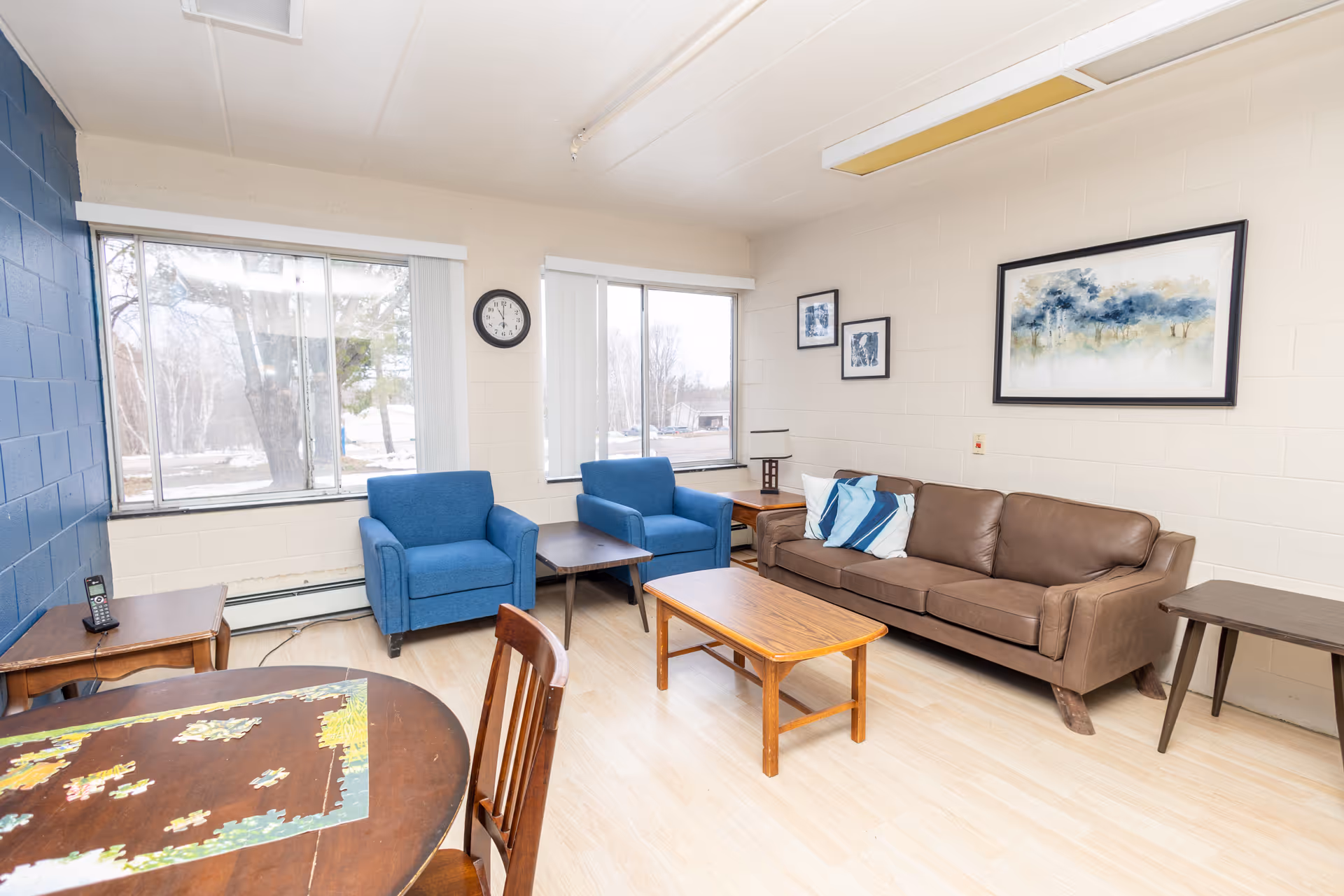 Bright common living room with two blue armchairs, a brown sofa, wooden tables, and large windows.