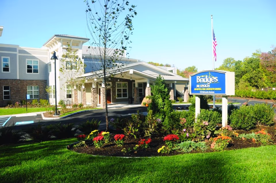 Exterior view of Bridges by EPOCH at Norwalk senior living facility with a landscaped garden featuring colorful flowers in the foreground, a driveway leading to the building entrance, and an American flag flying near the facility sign.
