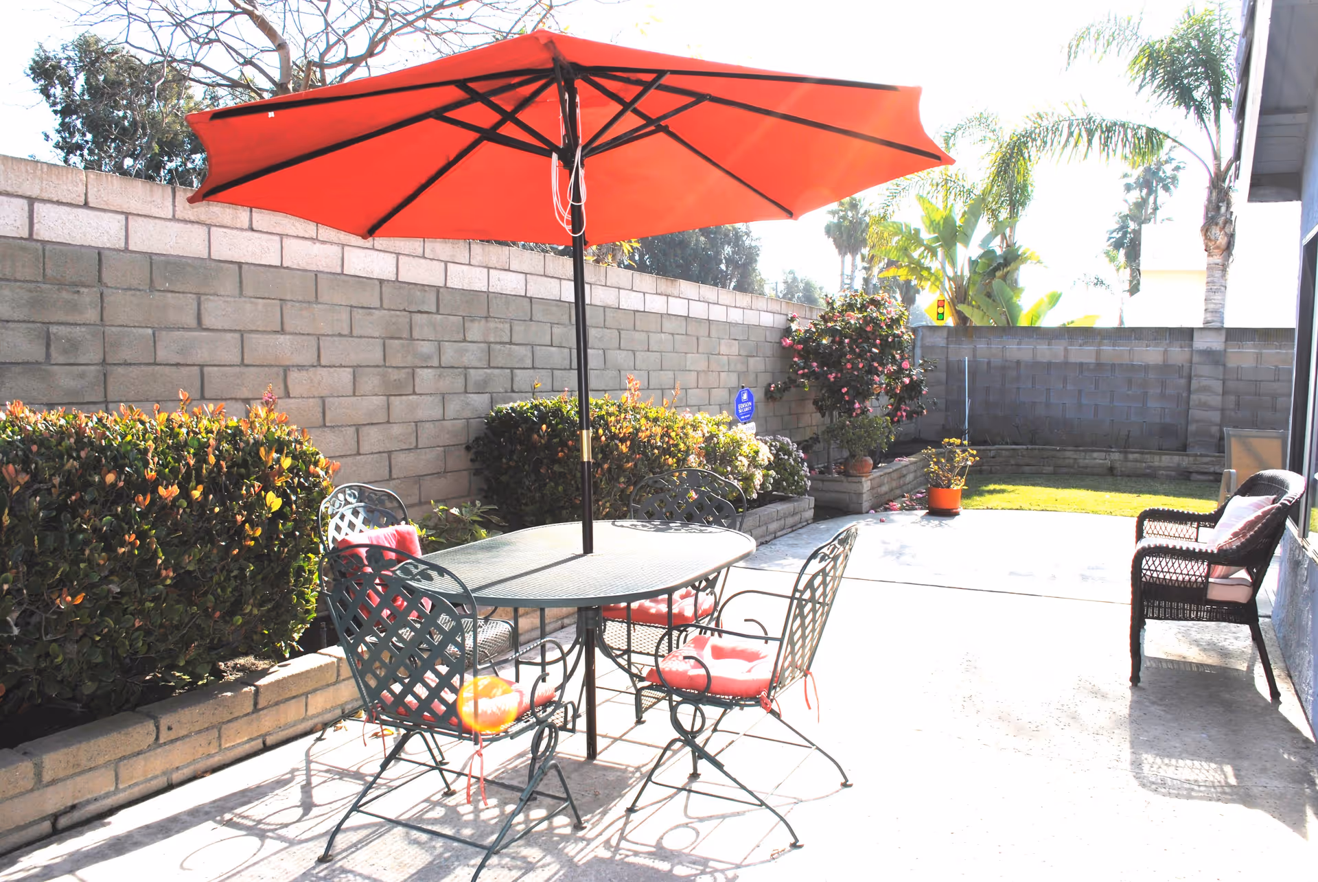 Outdoor patio area with a round metal table and four chairs with red cushions under a large red umbrella. There are bushes and potted plants along a brick wall, and a wicker bench with cushions is placed against the building on the right side. Palm trees and other greenery are visible in the background.