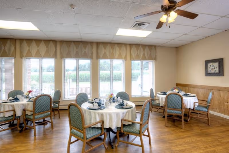 A bright dining room with round tables covered in white tablecloths, each set with plates, cups, and napkins. The room has wooden floors, several windows with blinds and valances, and a ceiling fan with lights. There are cushioned wooden chairs around each table and a framed picture on the wall.