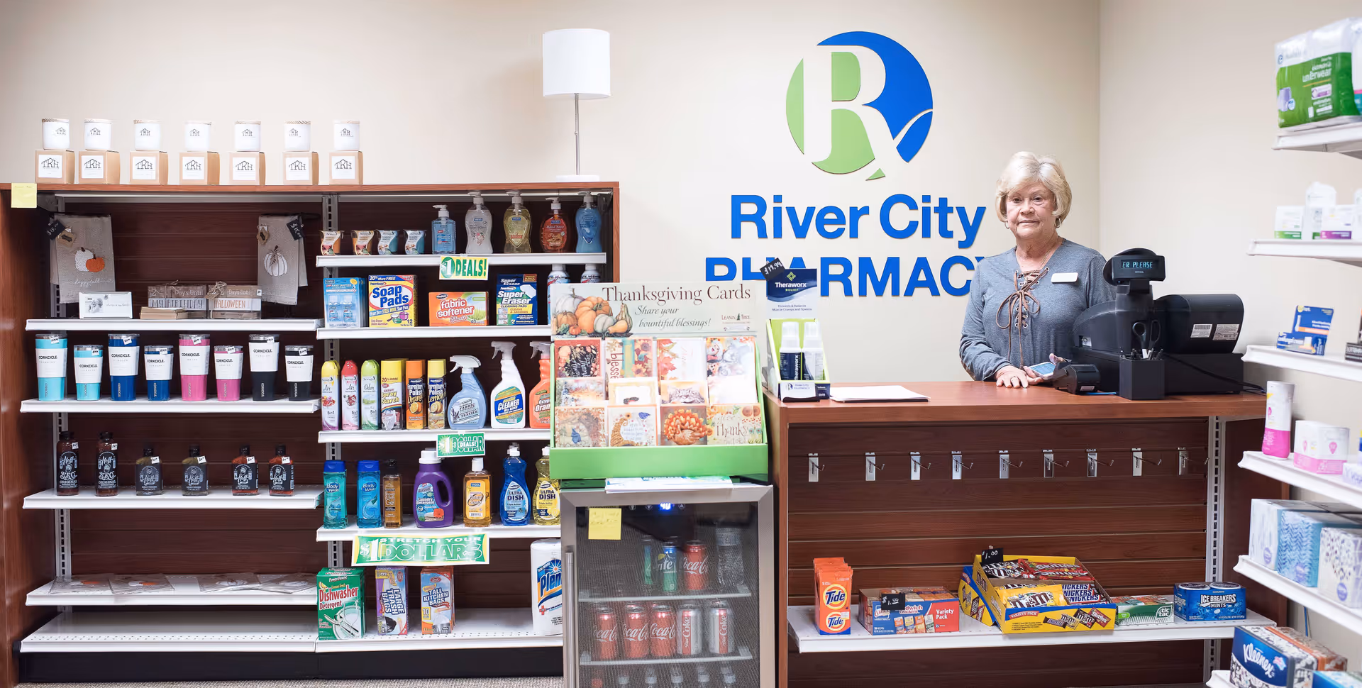 Interior view of a small pharmacy counter with a woman standing behind it. Shelves on the left and right display various household and personal care products. A refrigerator with beverages is in front of the counter. The wall behind the counter has a sign that reads 'River City Pharmacy.'