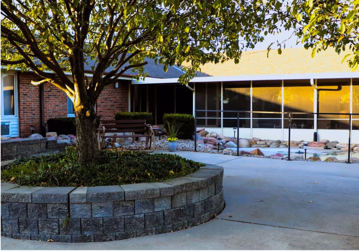 Courtyard with a tree in a raised stone planter, a bench, and a building with a screened porch in the background.