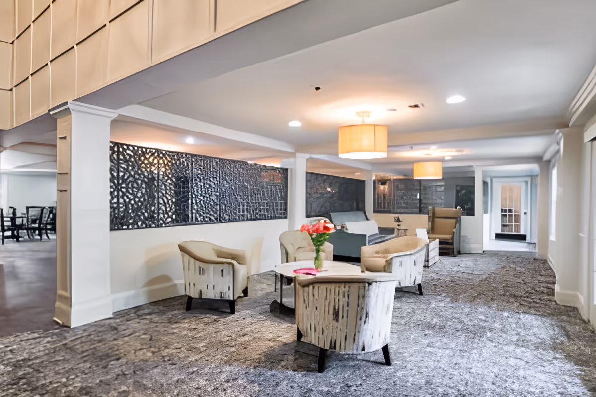 Carpeted communal lounge with upholstered chairs and sofas arranged around low tables, pendant lights, and a decorative wall panel.