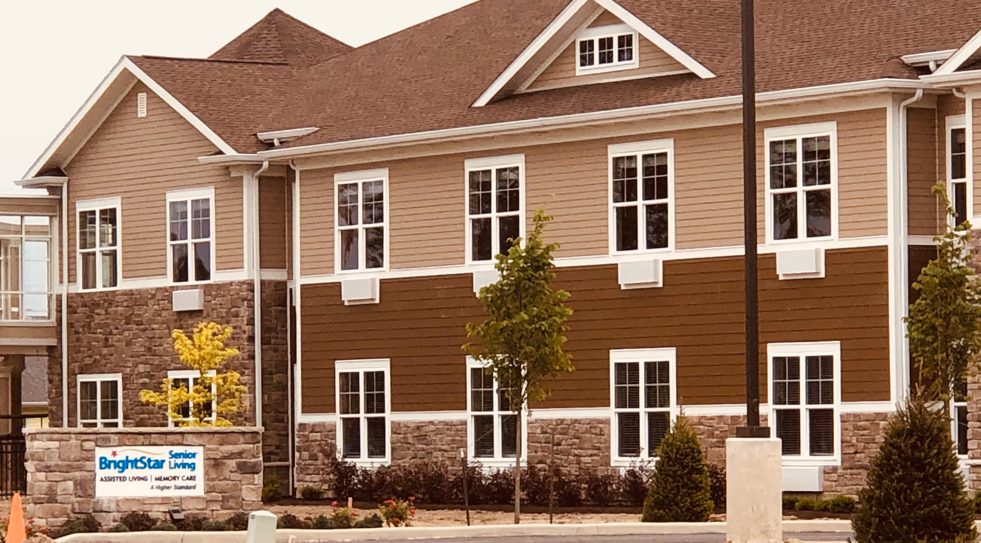 Exterior view of a two-story senior living facility building with beige and brown siding, multiple white-framed windows, and a stone base. There are small trees and shrubs planted in front of the building, and a sign that reads 'BrightStar Senior Living ASSISTED LIVING | MEMORY CARE A Higher Standard' is visible near the entrance.