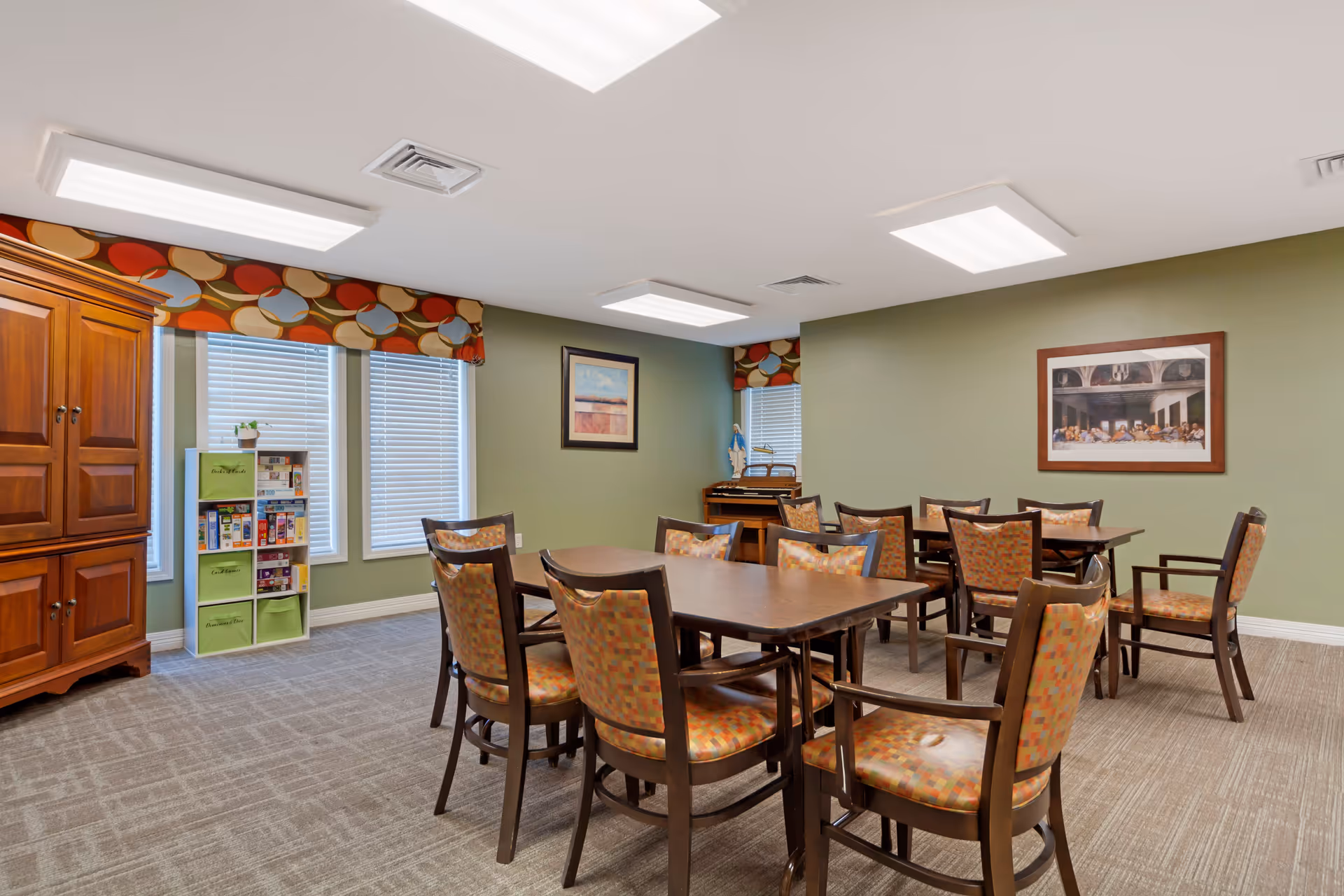 A well-lit room with two rectangular wooden tables surrounded by cushioned chairs with patterned upholstery. The walls are painted green and decorated with framed artwork. There is a wooden cabinet and a white shelving unit with board games near two windows with blinds and colorful valances. The room has a carpeted floor and ceiling lights.