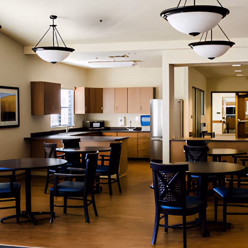 Interior view of a dining area with several round tables and chairs, adjacent to a kitchen with wooden cabinets, a refrigerator, a microwave, and a sink. The room has hanging ceiling lights and a framed picture on the wall.