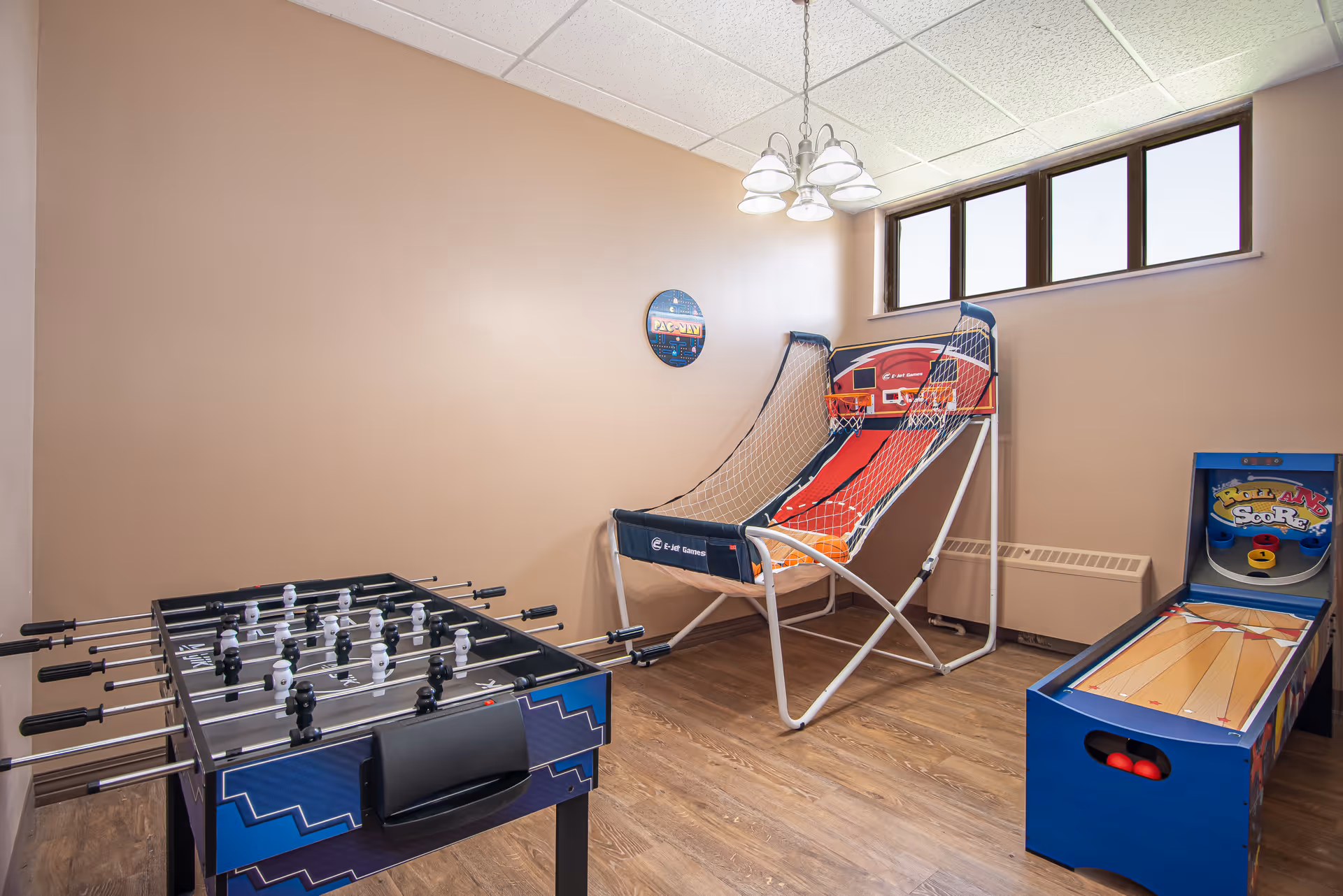 Small recreational room with a foosball table in the foreground, a basketball arcade game and a skee-ball style game against beige walls and a high window.