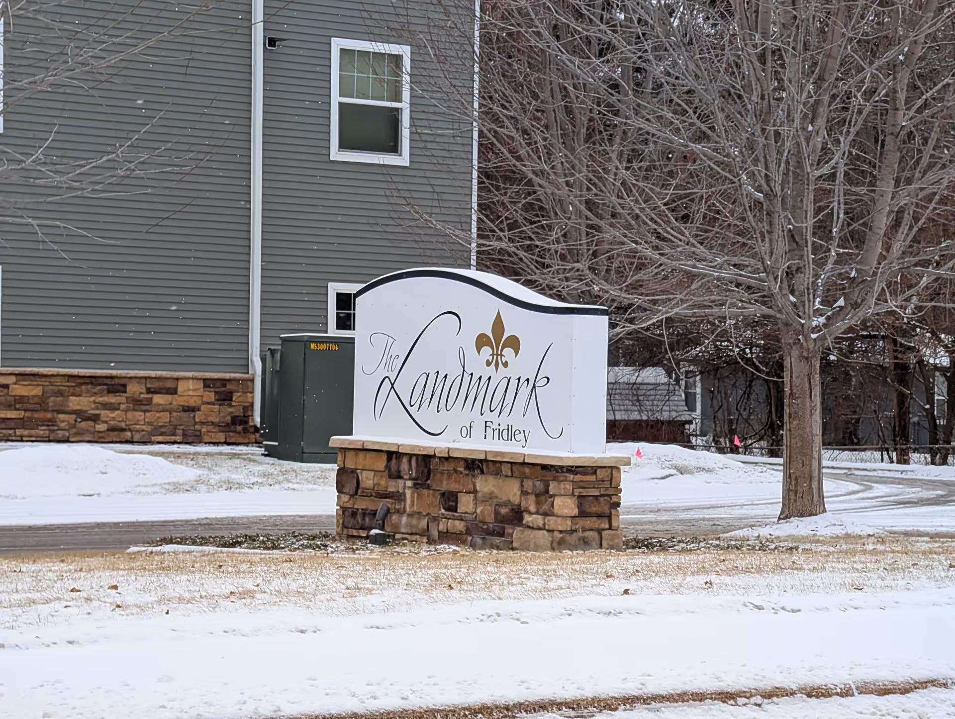 Outdoor view of a sign for The Landmark of Fridley, placed on a stone base with a building and leafless trees in the background during winter with snow on the ground.