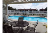 Outdoor patio area with a round glass table and four chairs under a pergola, overlooking a rectangular swimming pool surrounded by a white fence and residential buildings in the background.