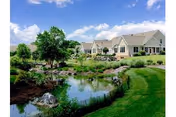 Exterior view of a senior living facility building with landscaped grounds, a pond, and a manicured lawn under a blue sky.