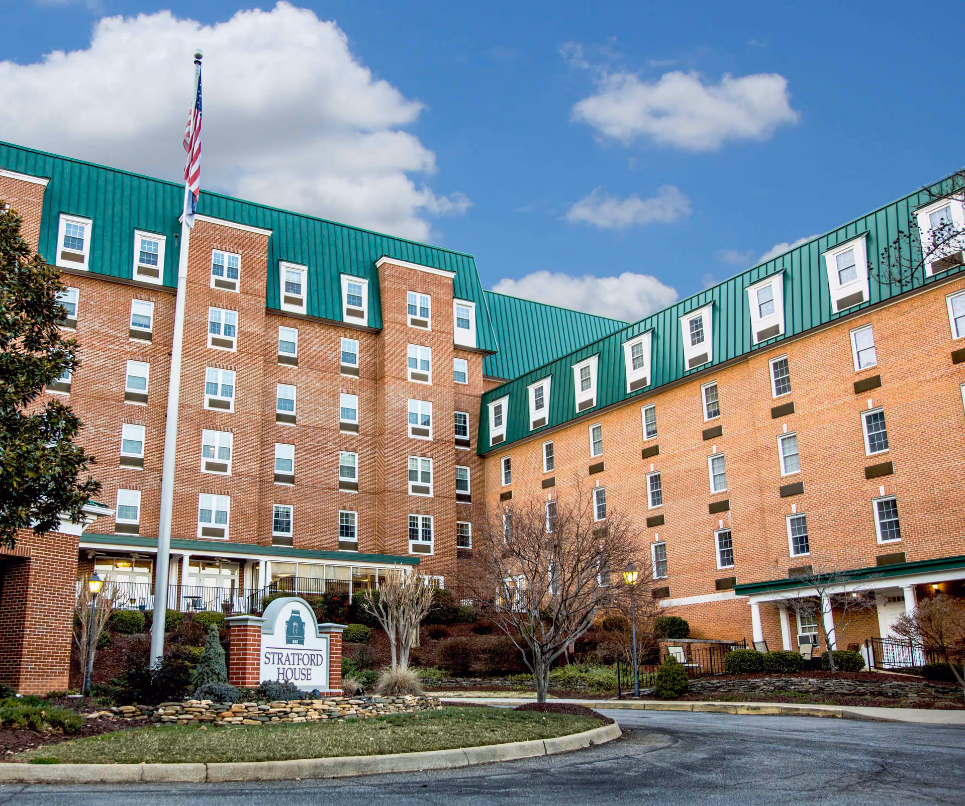 Brick multi-story senior living building with green mansard roof, flagpole, landscaping and a Stratford House entrance sign.