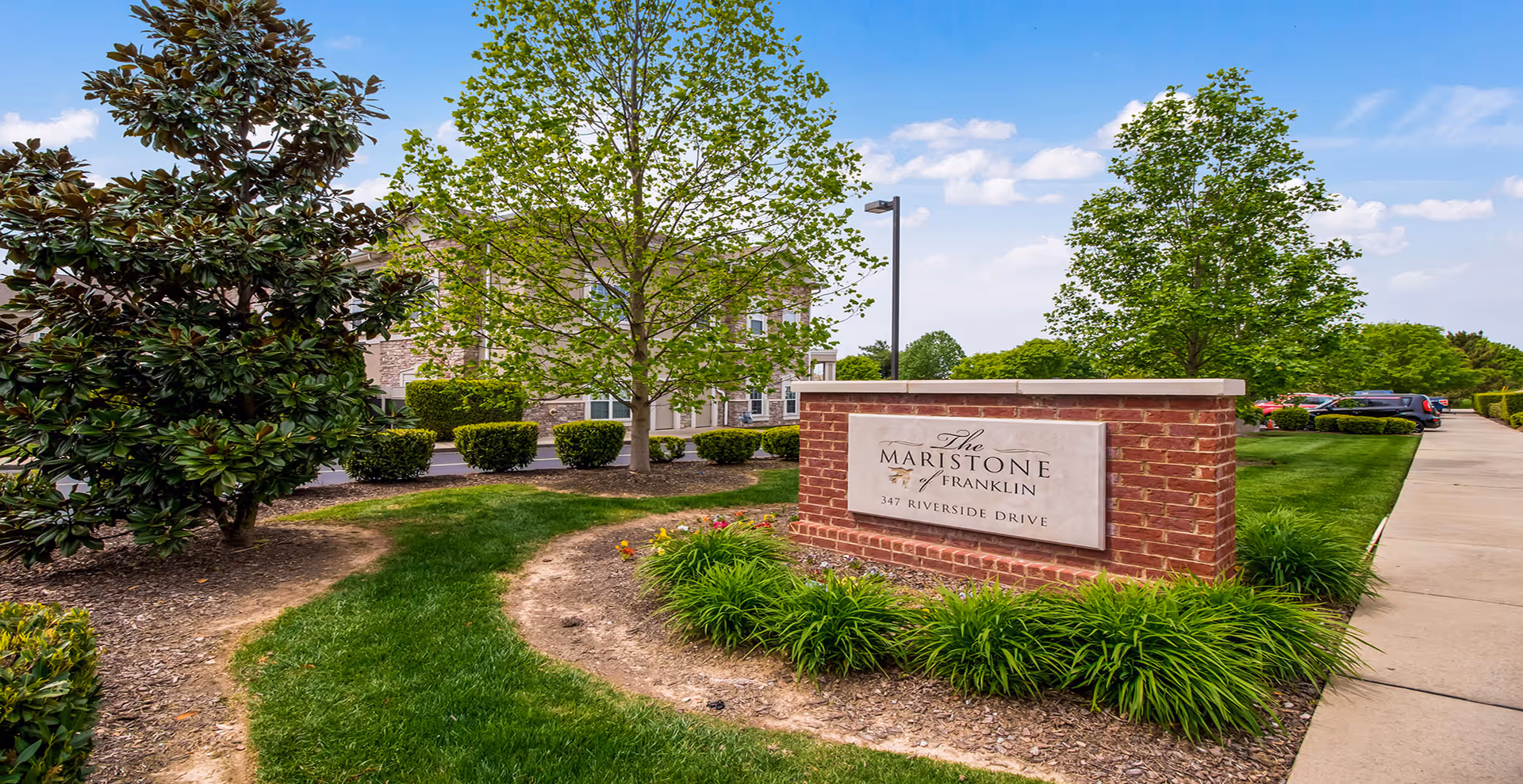 Outdoor view of the entrance area of The Maristone of Franklin facility, featuring a brick sign with the facility name and address, surrounded by green grass, trees, and shrubs, with a sidewalk and parked cars visible in the background under a partly cloudy sky.