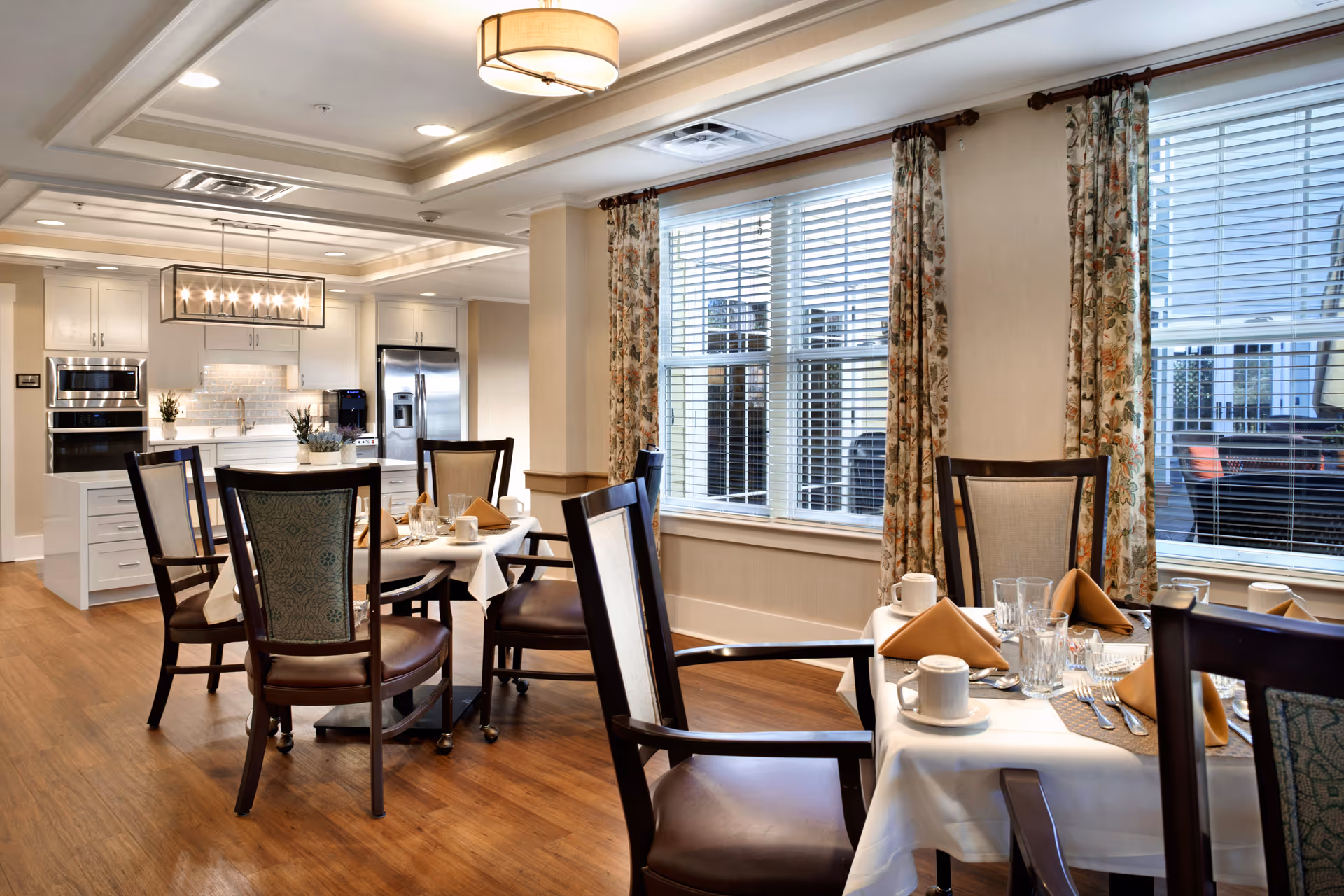 A bright dining room with wooden floors and several tables set with white tablecloths, beige napkins, glassware, and coffee cups. The room has large windows with floral curtains and white blinds, and a modern kitchen with white cabinets, stainless steel appliances, and a hanging light fixture is visible in the background.