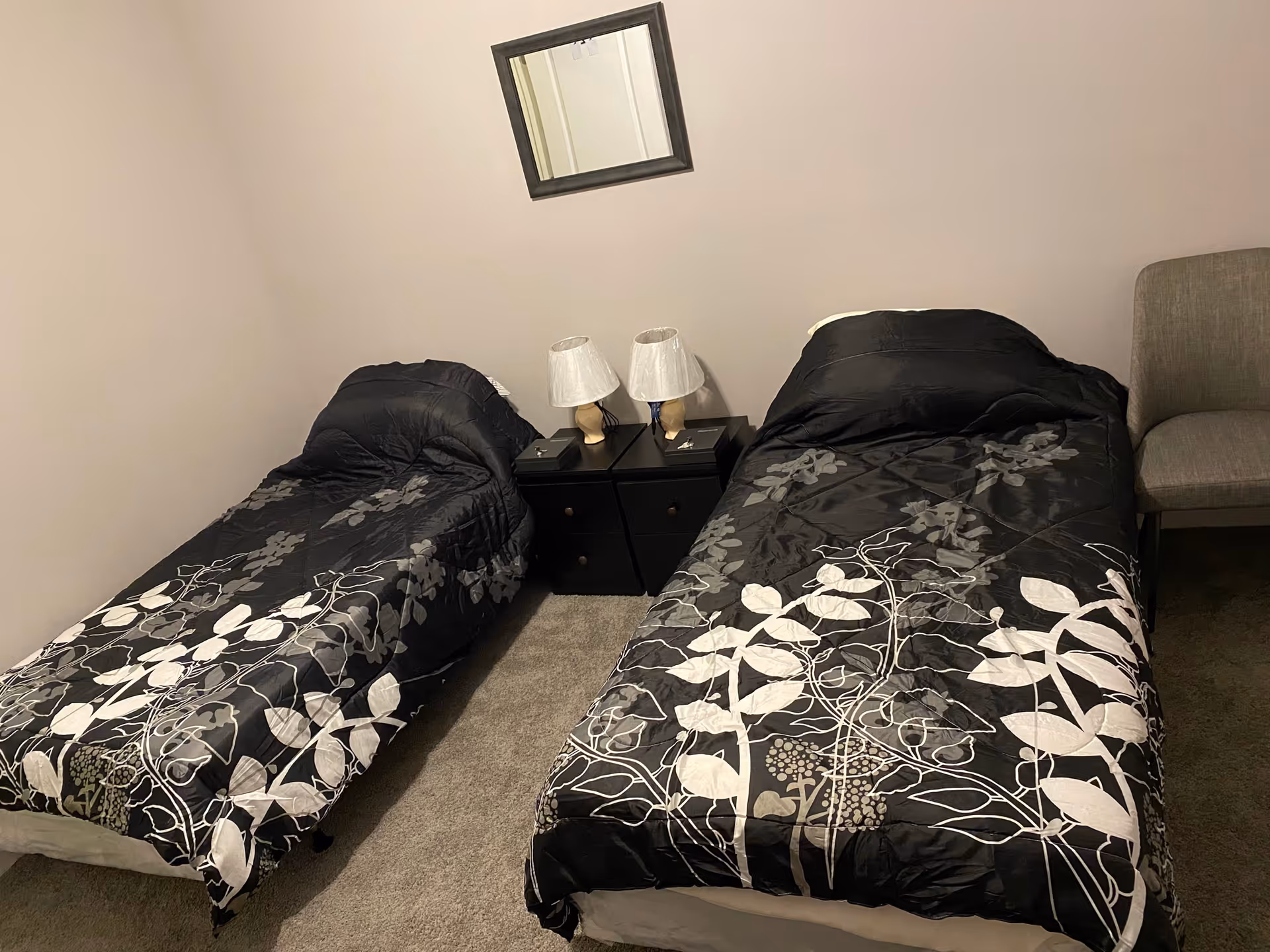 A small bedroom with two single beds covered in black and white floral patterned comforters. Between the beds is a black nightstand with two lamps and some small items. A gray upholstered chair is positioned to the right of the right bed. A small square mirror hangs on the beige wall above the nightstand.