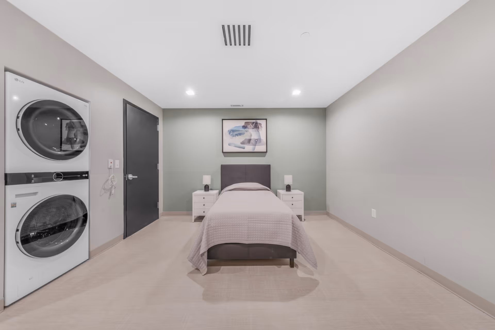 A simple bedroom with a single bed centered against a green accent wall. The bed has a gray headboard and is covered with a light gray quilt. On either side of the bed are white nightstands with small lamps. To the left of the bed, there is a black door and a stacked washer and dryer unit. The room has beige flooring and light gray walls, with recessed ceiling lights providing illumination.