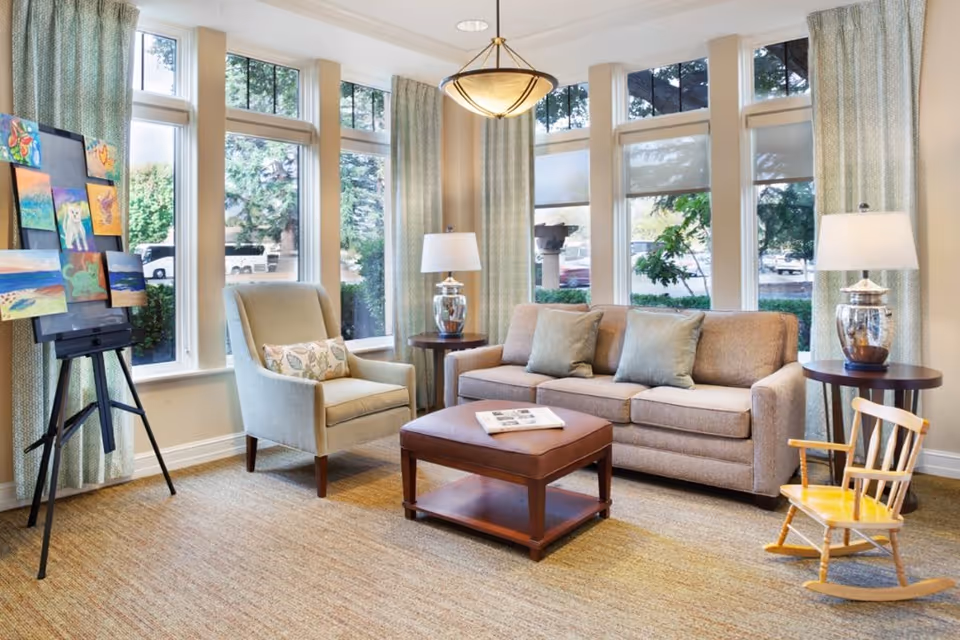 A cozy living room area with a beige sofa adorned with three cushions, a matching armchair with a decorative pillow, a wooden coffee table with a magazine on top, and a small wooden rocking chair. Large windows with light green curtains allow natural light to fill the room. An easel displaying colorful paintings is positioned near the windows, and two table lamps with glass bases sit on round wooden side tables beside the sofa.