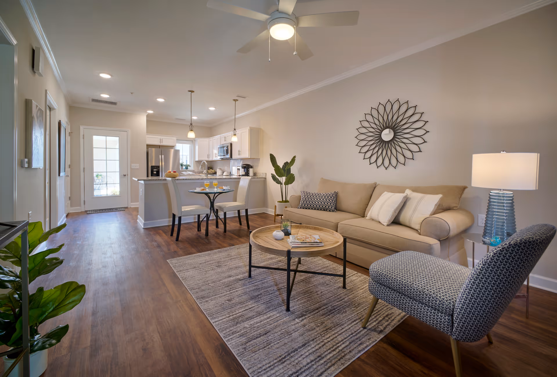 A modern, open-concept living room and kitchen area with wooden flooring. The living room features a beige sofa with pillows, a round wooden coffee table on a gray rug, a patterned armchair, a side table with a lamp, and a decorative wall mirror. The kitchen has white cabinets, stainless steel appliances, a small dining table with two chairs, and pendant lights hanging above the kitchen island.