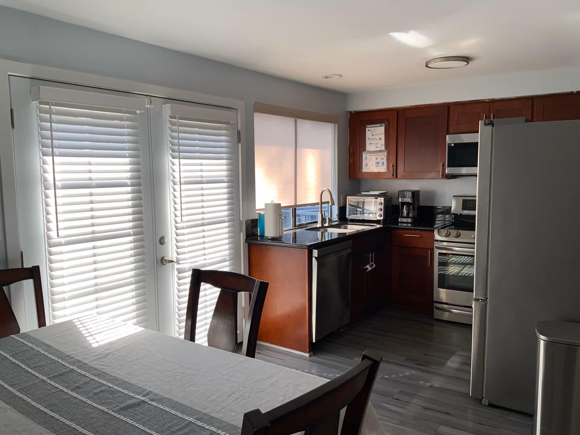 A modern kitchen with dark wooden cabinets, stainless steel appliances including a refrigerator, oven, microwave, and dishwasher. There is a double sink under a window with a roller shade, a toaster oven, and a coffee maker on the countertop. In the foreground, there is a dining table with a light-colored tablecloth and wooden chairs. French doors with white blinds allow natural light into the room.