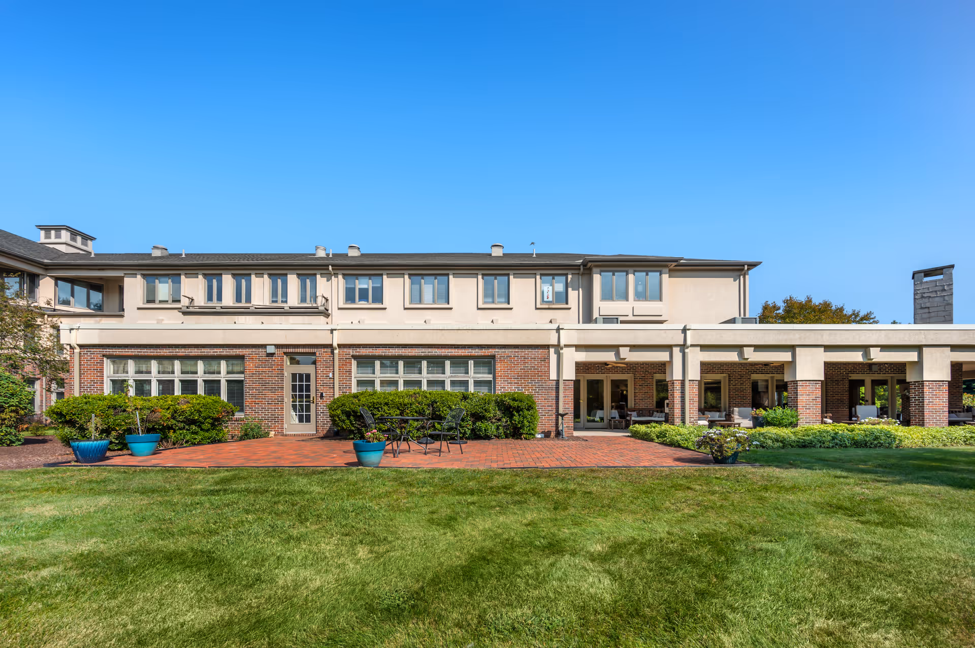 Exterior view of a two-story senior living facility building with a brick and beige facade, large windows, a patio area with outdoor seating, potted plants, and a well-maintained green lawn under a clear blue sky.