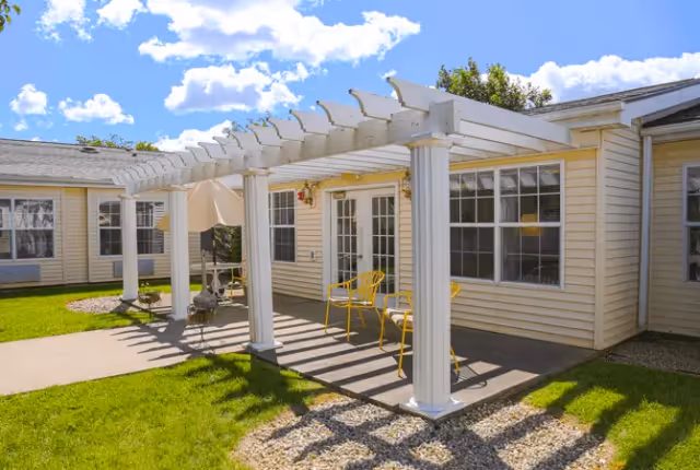 Outdoor patio area at a senior living facility with a white pergola supported by columns, yellow metal chairs, a table with an umbrella, and a beige building with multiple windows in the background under a blue sky with scattered clouds.
